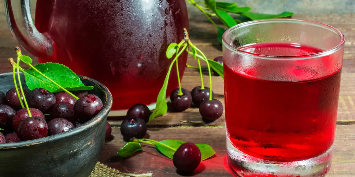 cold cherry juice in a glass and pitcher on wooden table with ripe berries in pottery bowl standing on canvas